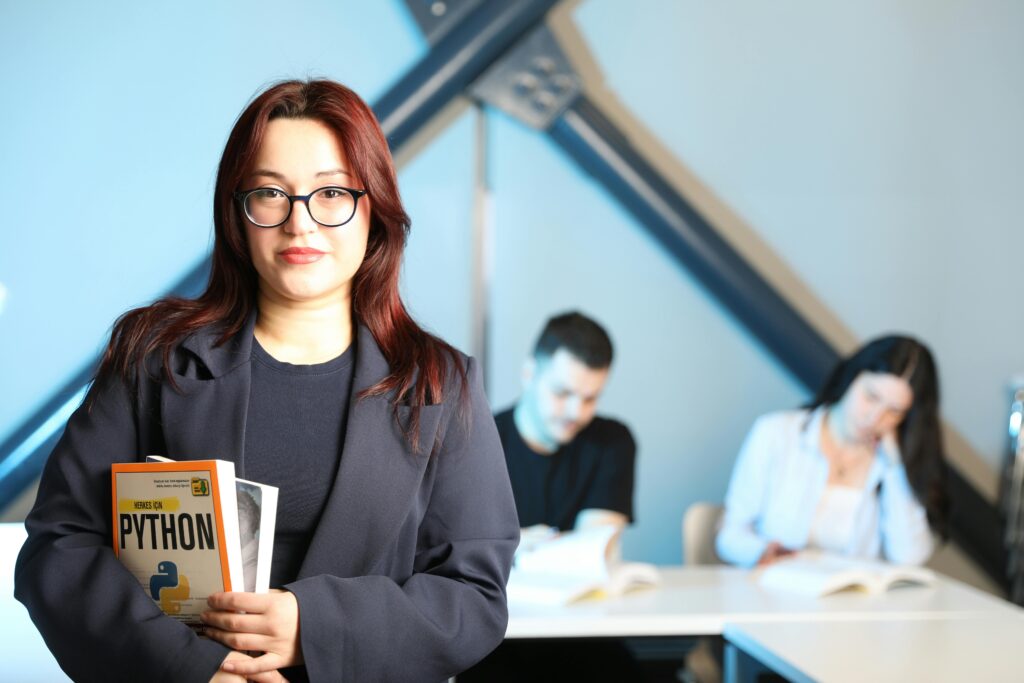 pexels-photo-32213227-32213227 Woman in classroom setting holding Python programming book, with students in background.